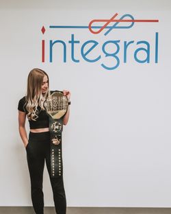 Kaitlyn holding the Integral graduation belt in front of a wall with the Integral logo on it
